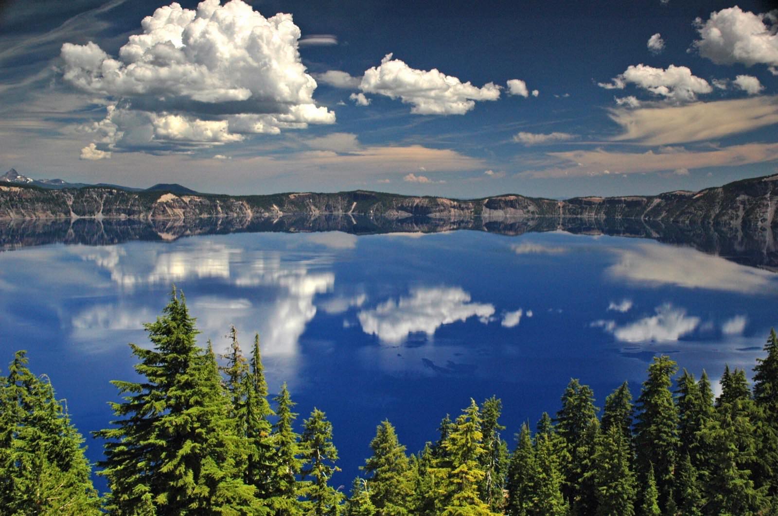 Crater Lake's deep blue caldera water reflecting clouds and the surrounding rim, with conifer trees in the foreground, Klamath County, Oregon.