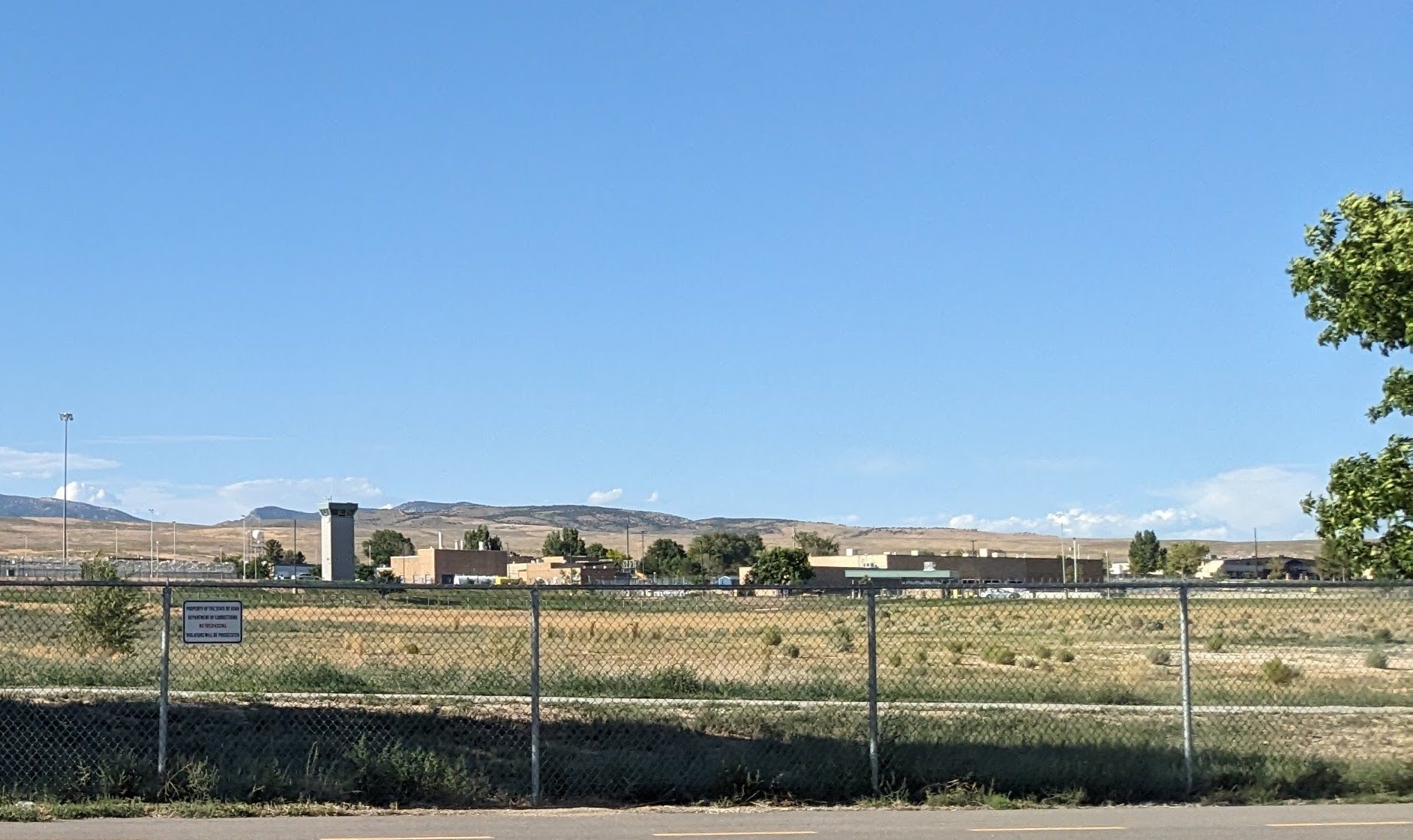A low prison complex with a guard tower stands behind a chain-link fence, with mountains in the distance.