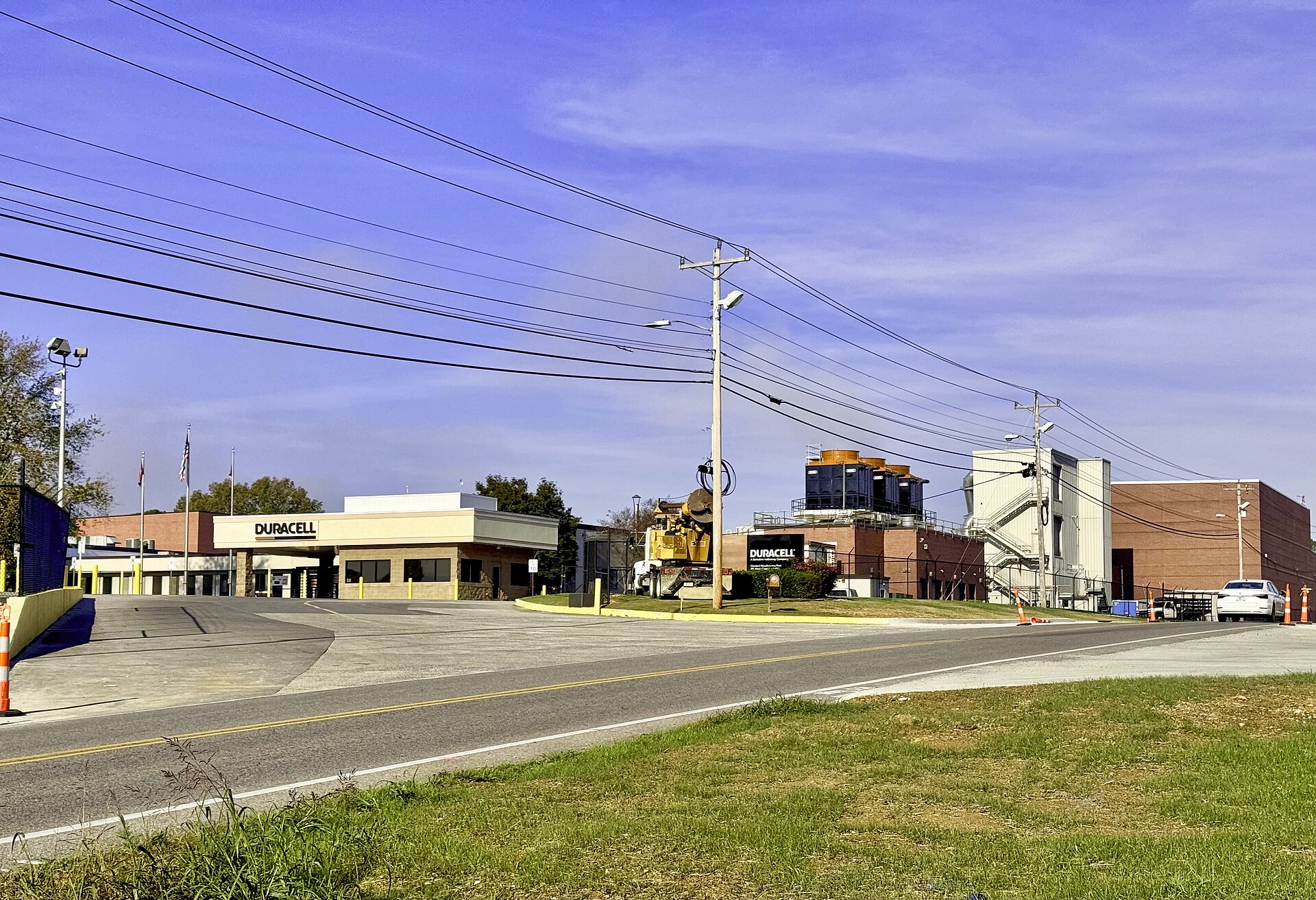 An industrial battery factory with signage, cooling equipment, and low buildings seen from the roadside.